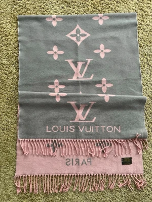 Louis Vuitton scarf i rosa och grått - Lyxig scarf från Louis Vuitton i mjuk mix av siden och ull. Dubbelsidig design i ljusrosa och grått med ikoniska LV-loggor och monogramblommor. Fransar längs kanterna ger en extra touch. Perfekt accessoar för att lyfta din outfit.