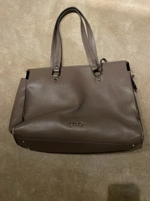 Beige handväska från Liu Jo - Snygg beige handväska från Liu Jo i strukturerat skinnimitation. Väskan har dubbla handtag, metallfärgade detaljer och en klassisk, stilren form. Perfekt för dig som vill ha en rymlig och trendig väska med exklusiv känsla.