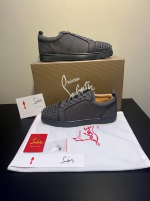 Christian Louboutin Skor - Säljer ett par gråa sneakers från Christian Louboutin i mocka med coola nitar på tån och klassisk röd sula. Skorna har snörning och en stilren, rund tå. Insidan är i beige skinn och de kommer med originalkartong och dustbag. Priset är diskuterbart vid snabb affär
