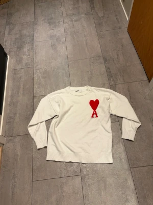 Vit stickad tröja från Ami - Säljer en vit stickad tröja från Ami Alexandre Mattiussi med röd hjärt- och A-detalj på bröstet. Tröjan har rund hals, ribbade muddar och lång ärm. Perfekt för dig som vill ha en clean men ändå statement look.