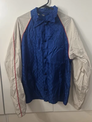 Gucci GG windbreaker jacket - Säljer en riktigt snygg blå och vit vindjacka från Gucci med diskret GG-monogram över hela jackan. Jackan har vita ärmar med röda detaljer längs sömmarna och knäppning framtill. Materialet är lätt och glansigt.