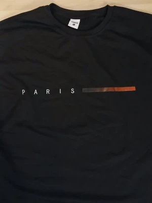 Svart långärmad t-shirt Paris Trend - Svart långärmad t-shirt från Trend i storlek XL. Framsidan har ett stilrent tryck med texten 'PARIS' och en färgad linje. Ärmarna är vita från armbågen och ner, vilket ger en cool kontrast. Tillverkad i 100% polyester för en lätt och skön känsla.