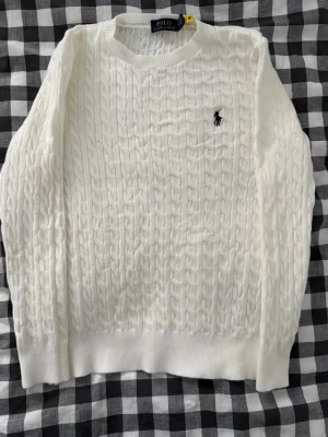 Vit kabelstickad tröja Polo Ralph Lauren - Kabelstickad vit tröja Polo Ralph Lauren med klassisk rund halsringning och broderad logga på bröstet. Tröjan har lång ärm och ribbade muddar. Perfekt för dig som gillar clean och tidlös stil. 😊👍