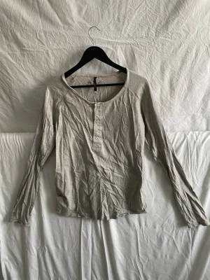 Ljus sopgrå långärmad topp med knappar - En enkel och stilren ljusgrå långärmad topp från nudie jeans co. Toppen har rund halsringning och twista knappar framtill som detalj. Tillverkad i mjuk bomull för en skön känsla. Perfekt till successiva lager eller för en chill look.