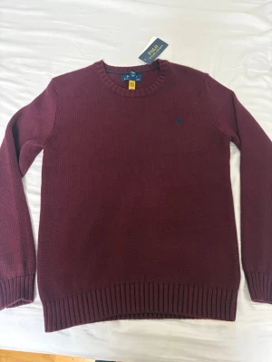 Vinröd stickad tröja Polo Ralph Lauren - Säljer en vinröd stickad tröja från Polo Ralph Lauren med klassisk rund hals och diskret blå logga broderad på bröstet. Tröjan har ribbade muddar vid ärmslut och nederkant, perfekt för dig som gillar stilrena och tidlösa plagg. Tröjan passade ej mig och försent för att lämna tillbaka, hör av dig om du är intresserad. Priset är ej hugget i sten!