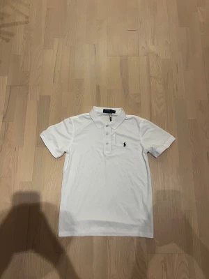 Vit piké från Polo Ralph Lauren - Klassisk vit pikétröja från Polo Ralph Lauren med korta ärmar och krage. Tröjan har tre knappar framtill och den ikoniska lilla loggan broderad på bröstet. Tillverkad i mjuk bomull som känns skön mot huden. Perfekt till jeans eller shorts.
