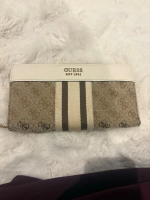 Beige och vit plånbok från Guess - Säljer en snygg beige och vit plånbok från Guess med klassiskt logomönster och två breda ränder framtill. Plånboken har dragkedja runtom, flera fack för kort och myntfack.