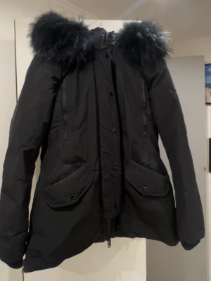 Svart Rockandblue parkas med päls - Säljer en svart parkas från Rockandblue i storlek 34. Jackan har stor, fluffig päls runt huvan, dragkedja och knappar framtill samt två stora fickor med lock. Perfekt för kalla dagar och riktigt snygg till vintern.