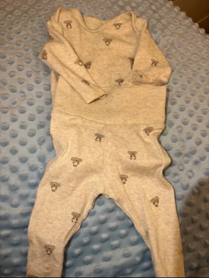 Gråmelerad babyset med nallemönster - Sött tvådelat babyset i gråmelerad bomull med små nallemönster över hela plagget. Setet består av långärmad tröja och byxor med mjuk resår i midjan. Perfekt för chill dagar hemma eller när du vill ha något gulligt och bekvämt.  Storlek 62 