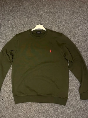 Mörkgrön sweatshirt från Polo Ralph Lauren - Säljer en mörkgrön sweatshirt från Polo Ralph Lauren med klassisk rund halsringning och röd broderad logga på bröstet. Tröjan har långa ärmar och ribbade muddar vid ärmslut och nederkant. Perfekt för en clean och stilren look.