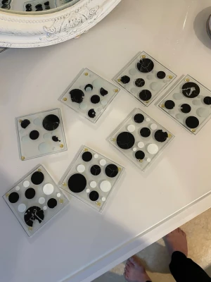 Svartvita glasunderlägg med prickar - Säljer ett set med sex fyrkantiga glasunderlägg i glas med svartvita prickar i olika storlekar. Underläggen har en modern och lekfull design med genomskinlig botten och små gula gummifötter i hörnen. Perfekt för att liva upp fikabordet eller skrivbordet.vissa av dem är färgen är av 