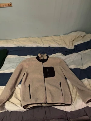 Beige teddyjacka från Sail Racing - Säljer en beige teddyjacka från Sail Racing med svart bröstficka och dragkedjor. Jackan har hög krage, hel dragkedja framtill och två sidofickor med dragkedja. Perfekt för kyliga dagar och riktigt skön att ha på sig.