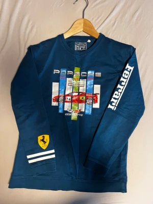Blå långärmad tröja med biltryck - Blå långärmad tröja med tryck av sportbil och texten Porsche, Corvette och Ferrari. Stor Ferrari-logga på ena ärmen och vita ränder vid ärmslutet. Perfekt för dig som gillar motorsport och snygga prints.