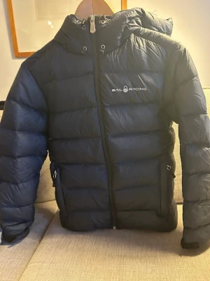 Svart pufferjack mill Sail Racing - Säljer en svart pufferjacka från Sail Racing medactivities logga på bröstet och huvan. Jackan har huva, dragkedja framtill och två sidofickor med dragkedja. Insidan är grå och materialet känns lätt och smidigt. Perfekt för kalla dagar när du vill hålla värmen. Lite skynken men med vädring är det som en ny jacka. Inga defekter eller hål 