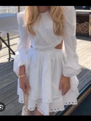 Vit kortklänning med volang och öppen rygg - Superfin vit kortklänning med långa puffärmar, volangig kjol och broderade detaljer längs kanterna. Klänningen har snygga cut-outs i midjan och öppen rygg som knäpps upptill. Perfekt för studenten och sommaren, klänning är från bik bok 