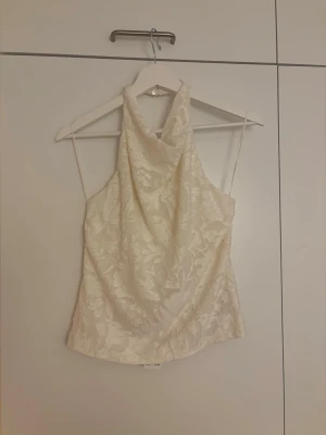 Krämvit halterneck topp med blommönster - Snygg halterneck topp från Gina Tricot i krämvitt med subtilt blommönster. Toppen har en draperad framsida och är ärmlös. Perfekt för dig som vill ha en elegant och trendig look.