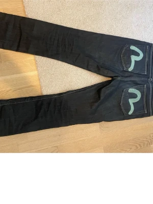 Svarta Evisu jeans med gröna detaljer - Säljer ett par svarta jeans från Evisu med  ikoniska gröna sömmar och på bakfickorna. Jeansen har klassisk femficksdesign, kontrastsömmar och coolt mönstrat innerfoder. Perfekt för dig som gillar street och vill sticka ut.