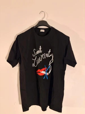 Svart Saint Laurent t-shirt med tryck - Svart t-shirt från Saint Laurent med coolt grafiskt tryck framtill i vitt, rött och blått. Motiv med texten 'Saint Laurent', röda läppar och blå detaljer. Klassisk passform och rund hals. Perfekt för dig som vill sticka ut med en ikonisk designertee. Axelmåttet är 50cm och den är ungefär 68cm lång. Storlek L men kan möjligtvis passa M med.