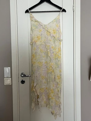 Blommig långklänning från H&M - Säljer en romantisk långklänning från H&M med blommigt mönster i ljusa toner av gul, lila och grön. Klänningen har volangdetaljer, smala axelband och asymmetrisk nederkant. Perfekt för sommarens alla tillfällen! (Den är skrynklig då den legat undan packad med sommarkläder under vintern)