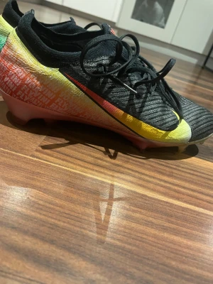 Nike fotbollsskor Flyknit svart/röd - Säljer ett par Nike fotbollsskor, ena skon är svart med gul och röd gradient och den andra är röd med ljusblå swoosh. Skorna har snörning och är tillverkade i lätt syntetmaterial med Flyknit-teknologi för extra komfort och passform. Perfekta för dig som vill sticka ut på planen. Svarta köpta från unisport elite som är Vini jr skor har box till dem, de röda skorna är PRO från unisport man får med båda på samma köp.