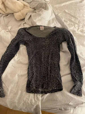 Leopardmönstrad topp från ONLY - Säljer en långärmad topp från ONLY i svart och grått med leopardmönster. Toppen har v-ringning och knappar framtill, samt är ribbad i materialet vilket ger en snygg och bekväm passform. Perfekt för dig som gillar djurmönstrat och vill sticka ut lite extra. Den har ej några defekter. 