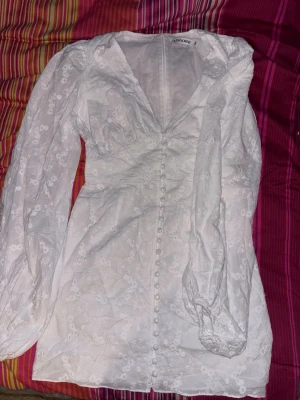 Vit blommig kortklänning från Adoore - Supersöt vit kortklänning studentklänning från Adoore med v-ringning, puffiga långärmar och knappar framtill. Klänningen har ett diskret blommigt mönster och är gjord i ett tunt, lätt material som känns perfekt för varmare dagar.