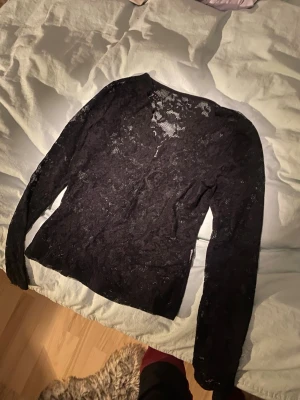Svart spetsblus med lång ärm - Säljer en svart blus i genomskinlig spets med blommigt mönster. Blusen har långa ärmar och en figurnära passform. Perfekt för dig som gillar detaljer och vill ha något extra till din outfit.