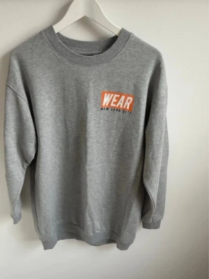 Grå sweatshirt från Gina Tricot - Grå sweatshirt från Gina Tricot med tryck 'WEAR New York City' i vitt och orange på bröstet. Klassisk rund halsringning och ribbade muddar vid ärmslut och nederkant. Perfekt för chill dagar och enkel att styla med jeans eller leggings.