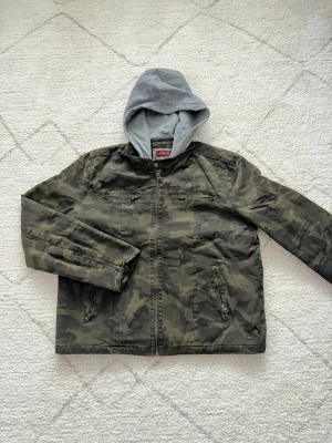 Levi's camo vindjacka med huva - Säljer en grön och grå kamouflagemönstrad vindjacka från Levi's med grå huva i sweatshirtmaterial. Jackan har dragkedja framtill, flera fickor med dragkedjor och en cool streetstil. Perfekt för dig som gillar en avslappnad look.