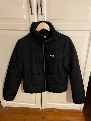 Svart pufferjacka från Vans - Säljer en svart pufferjacka från Vans med hög krage och dragkedja framtill. Jackan har en clean look med diskret Vans-logga på bröstet och två fickor på sidorna. Perfekt för kalla dagar och enkel att matcha med allt.