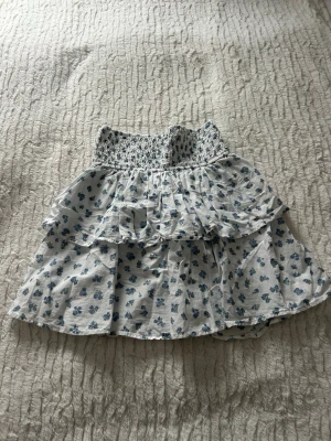 Vit blommig volangkjol i bomull - Supersöt kort kjol i vitt med blått blommönster och bred smock i midjan. Kjolen har dubbla volanger som ger en flowy känsla och är gjord i tunn bomull, perfekt för varma dagar. Passar dig som gillar romantisk och somrig stil.