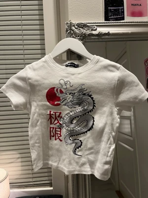 Vit t-shirt med draktryck  - Vit t-shirt i bomull  med coolt draktryck och röd sol på framsidan. T-shirten har korta ärmar, rund halsringning och röda japanska tecken. Perfekt för dig som vill sticka ut med en unik och trendig look.