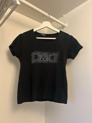 Svart D&G t-shirt med strass - Svart t-shirt från D&G med korta ärmar och rund halsringning. Framsidan har en stor D&G-logga i glittrande strass. Modellen är croppad och sitter snyggt på kroppen. Perfekt för dig som gillar en stilren men ändå blingig look. Köpt second hand så har ingen aning om den är äkta eller inte.