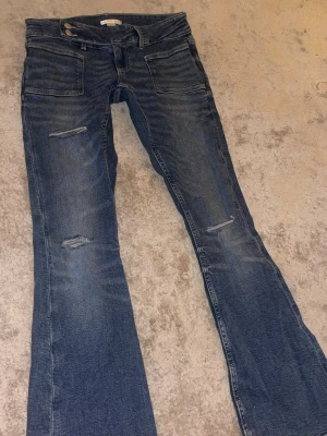 Blå bootcut jeans med slitningar - Säljer ett par blå jeans med bootcut passform och snygga slitningar på benen. Jeansen har stora fickor fram och klassisk knappgylf. Perfekta för dig som gillar en avslappnad men trendig stil. använda 1 gång