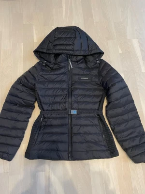 Svart pufferjacka från Calvin Klein S - Säljer en svart pufferjacka från Calvin Klein i storlek S. Jackan har huva, markerat midjebälte med metallspänne och dragkedja framtill. Quiltad design och två fickor med dragkedja. Perfekt för kalla dagar och riktigt snygg passform. 