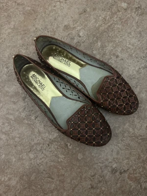 Bruna ballerinas från Michael Kors - Snygga bruna ballerinas från Michael Kors med utstansat mönster och guldfärgade nitar. Skorna har rund tå och är tillverkade i skinn med dekorativa detaljer över hela ovandelen. Perfekta för dig som vill ha något unikt och stilrent.