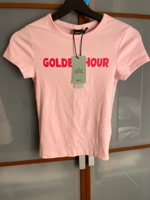 Rosa t-shirt Gina Tricot - Snygg ljusrosa t-shirt från Gina Tricot i mjuk bomull. Framsidan har trycket 'GOLDEN HOUR' i starkt röd färg. Modellen är kortärmad med rund hals och har en slim passform som sitter snyggt. Perfekt för dig som gillar trendiga statement-plagg.