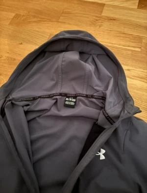Grå och svart vindjacka Under Armour S - Snygg tvåfärgad vindjacka från Under Armour i grått och svart. Jackan har huva med snörning, hel dragkedja framtill och en vertikal bröstficka med dragkedja. Materialet är lätt och känns sportigt, perfekt för utomhusaktiviteter. Under Armour-logga på bröstet.