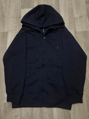 Marinblå zip-hoodie – Polo Ralph Lauren - Snygg och stilren marinblå zip-hoodie med klassisk röd Polo Ralph Lauren-logga på bröstet. Bekväm passform med dragkedja hela vägen, justerbara snören vid huvan och ribbade muddar.
