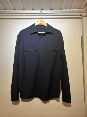 Mörkblå overshirt från Zara, M - Snygg mörkblå overshirt från Zara med klassisk krage och två stora bröstfickor med knappar. Jackan har långa ärmar och knäppning framtill. Perfekt för lager-på-lager och stilrena outfits. Materialet är mjukt och känns skönt mot huden. Jag har aldrig använt den så den är som ny. 