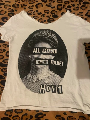 Vit Hov1 t-shirt med statementtryck - Säljer en vit t-shirt från Hov1 med ett stort svart statementtryck på framsidan. Trycket föreställer en kunglig person med texten 'All makt tillhör folket' i collage-stil. T-shirten har rund hals och korta ärmar, tillverkad i mjuk bomull. Köpt på vinted för 300. Den är avklippt så kan inte kolla storleken men skulle gissa på xs-s