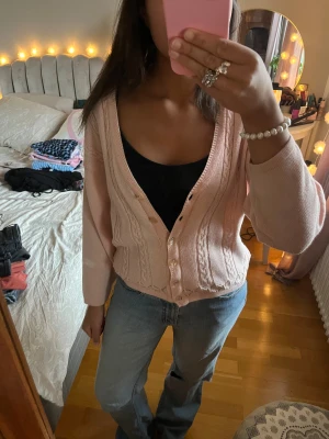 Ljusrosa stickad kofta med knappar - Superfin ljusrosa stickad kofta med snygga kabelstickade detaljer och pärlemorknappar framtill. Koftan har v-ringad hals och långärmade, lite loose ärmar.