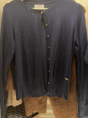 Mörkblå kofta från Holly & Whyte - Mörkblå kofta från Holly & Whyte med guldiga knappar framtill. Klassisk rund halsringning och ribbade muddar. Perfekt att slänga över en t-shirt eller klänning för en chill och stilren look.