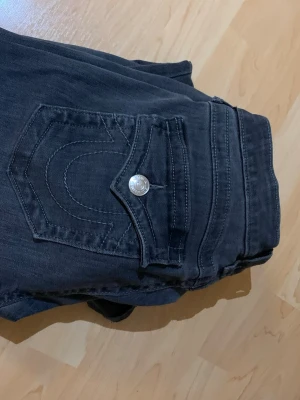 True Religion Becca Bootcut Jeans Mörkgrå - Svarta jeans från True Religion, modell Becca Mid Rise Bootcut. Snygga detaljer med markerade sömmar och ikonisk ficka med knapp bak. Bootcut-snitt och midwaist för en chill och klassisk look. Perfekta för dig som gillar street och premium denim. Säljer då de är för stora för mig