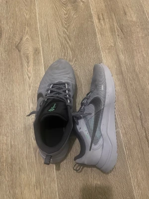 Grå Nike sneakers strl 41 - Säljer ett par grå Nike sneakers med svart swoosh och detaljer i grönt på sulan och plösen. Skorna har snörning och en rund tå, tillverkade i syntetmaterial och mesh för bra ventilation. Snygg och modern design som passar perfekt till streetwear.