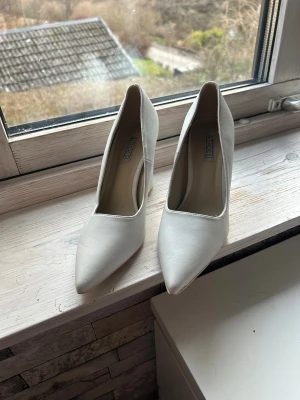 Vita pumps från NLY Shoes, storlek 37 - Snygga vita pumps från NLY Shoes i storlek 37. Skorna har spetsig tå och hög klack med en stilren, minimalistisk design. Tillverkade i syntetmaterial med slät yta och en stadig klack som ger extra höjd. Perfekta för dig som vill ha en clean och chic look. Använda en gång 