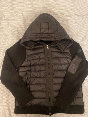 Svart Moncler pufferjacka med huva - Svart pufferjacka från Moncler med stickade ärmar och quiltad kropp. Jackan har huva, dragkedja framtill och diskret Moncler-logga på ärmen. Perfekt för kyliga dagar med sin kombination av dun och stickat material.