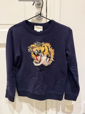  Gucci tröja med tigerbroderi - Snygg mörkblå tröja från Gucci med ett stort, detaljerat broderi av en rytande tiger på bröstet. Tröjan har rund hals, långa ärmar och är tillverkad i mjuk bomull. Perfekt för dig som vill sticka ut med en exklusiv och unik look.                                            Bara att höra av sog om det är något!