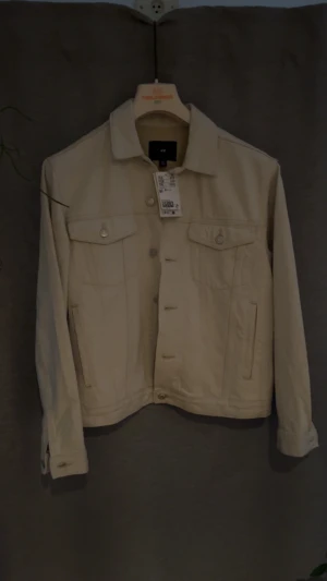 Beige jeansjacka från H&M - Snygg beige jeansjacka från H&M med klassisk krage, knappar framtill och två bröstfickor med lock. Jackan har långa ärmar med knapp vid mudden och raka sidofickor. Perfekt för dig som gillar stilrena och enkla plagg. Den är aldrig använd 