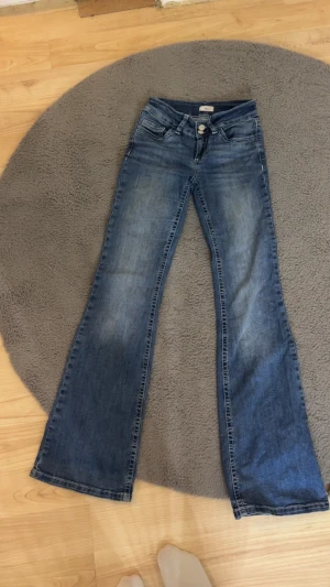 Blå bootcut jeans från nelly - Snygga blå jeans från Nelly med bootcut passform och klassisk femficksdesign. Jeansen har ljusare tvättade partier, broderad bakficka med vit vågig detalj och slitna kanter vid benslut. Materialet är robust denim som ger en cool vintagekänsla.